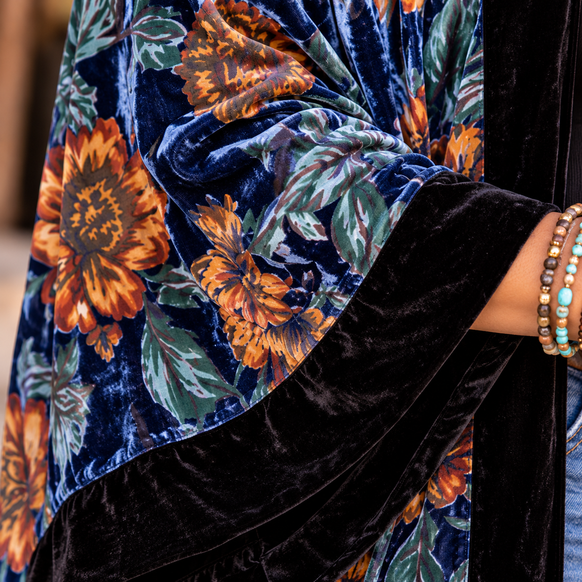 A close-up of a person wearing a navy blue velvet shawl with a large orange and green floral print and solid black velvet trim, accessorized with beaded bracelets.