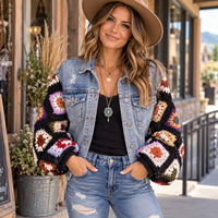 A woman wearing a light-wash denim jacket with colorful crocheted granny-square pattern sleeves, a black top, blue jeans, and a tan hat, standing on a storefront porch.