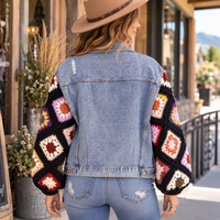 A woman viewed from behind wearing a light blue denim jacket with colorful crocheted granny-square sleeves, paired with light-wash jeans and a tan hat, standing on a sidewalk in front of a storefront.