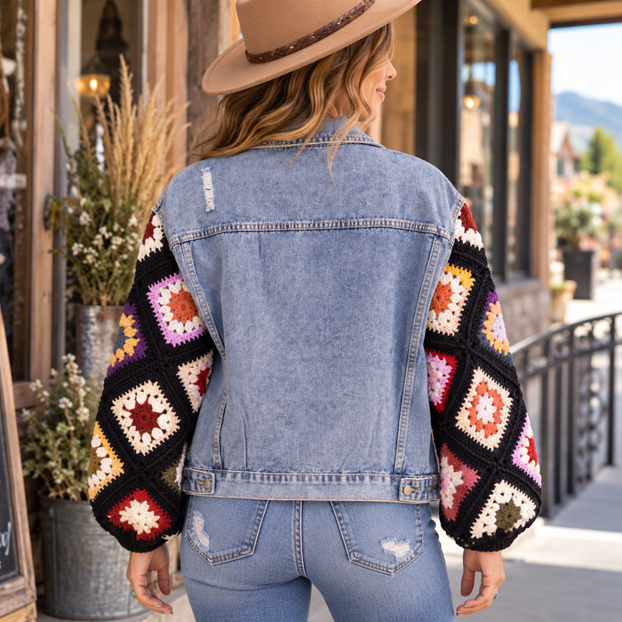 A woman viewed from behind wearing a light blue denim jacket with colorful crocheted granny-square sleeves, paired with light-wash jeans and a tan hat, standing on a sidewalk in front of a storefront.