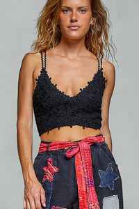 A woman modeling a black crochet lace camisole crop top with thin straps, paired with dark denim patchwork pants featuring a red patterned tie belt, against a solid light gray studio background.