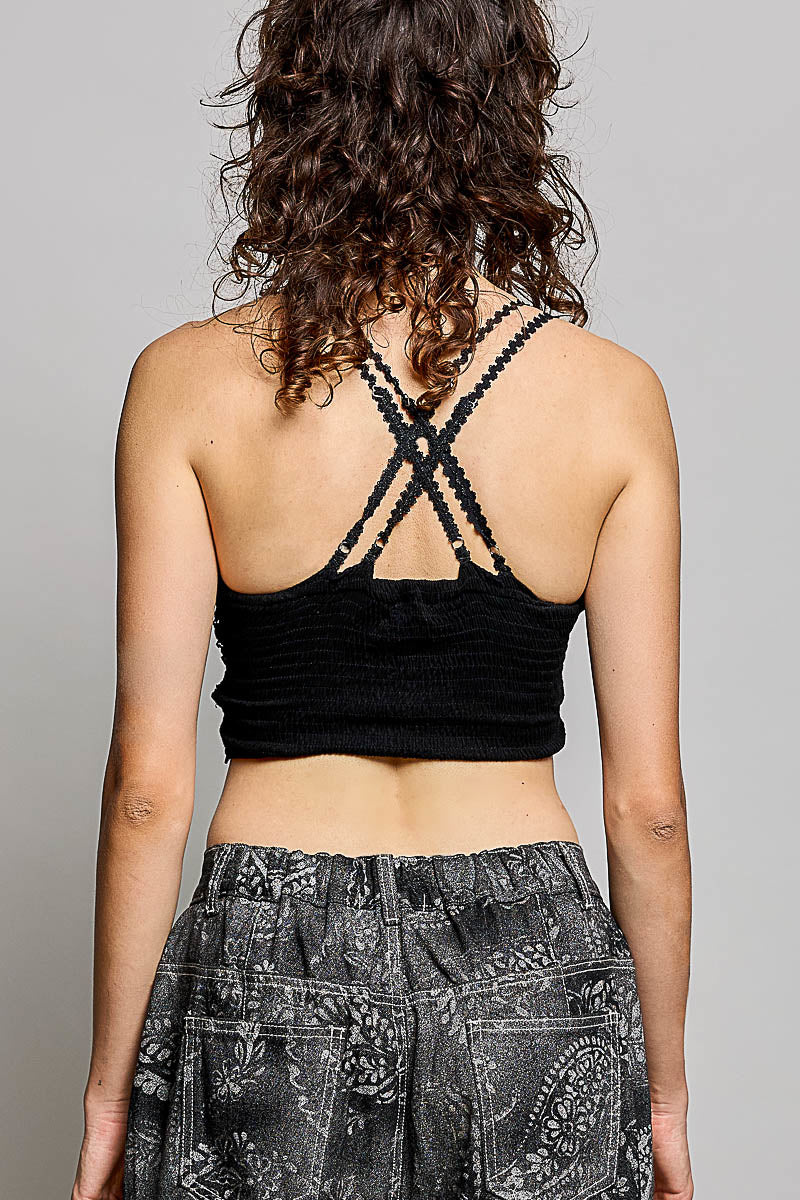 Rear view of a woman with curly brown hair wearing a black ribbed crop top with crisscross lace-up back straps and gray paisley patterned pants, against a light gray studio background.