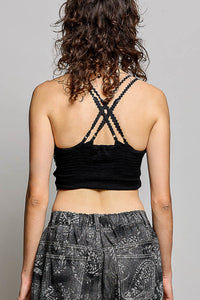 Rear view of a woman with curly brown hair wearing a black ribbed crop top with crisscross lace-up back straps and gray paisley patterned pants, against a light gray studio background.