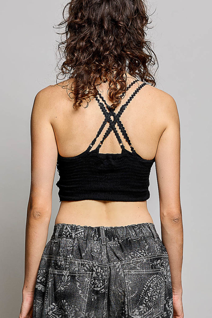 Rear view of a woman with curly brown hair wearing a black ribbed crop top with crisscross lace-up back straps and gray paisley patterned pants, against a light gray studio background.