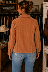 A woman viewed from behind wearing a rust-brown corduroy jacket and light blue jeans inside a rustic clothing boutique.
