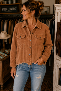 A woman wearing a brown corduroy button-up shirt jacket and distressed blue jeans, standing inside a rustic clothing boutique.