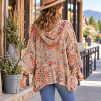A woman wearing a beige hooded patchwork poncho with red and orange floral prints, blue jeans, and a tan hat, photographed from behind on an outdoor storefront walkway.