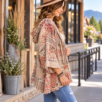 A woman wearing a beige hooded cardigan with a red and green floral patchwork print, blue jeans, and a wide-brim straw hat, standing on a sunny outdoor walkway next to a rustic wooden building.