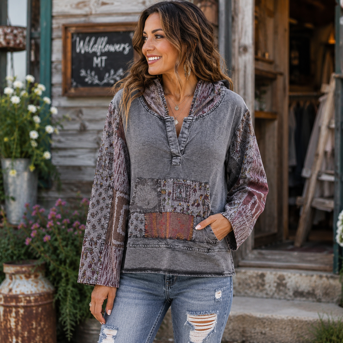 A woman wearing a gray patchwork hoodie sweatshirt with patterned sleeves and pockets, paired with distressed blue jeans, standing in front of a rustic wooden shop building.