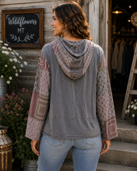 A woman viewed from behind wearing a gray hooded patchwork jacket with dusty pink and beige patterned sleeves, standing in front of a rustic wooden storefront with a chalkboard sign that reads 'Wildflowers MT'.