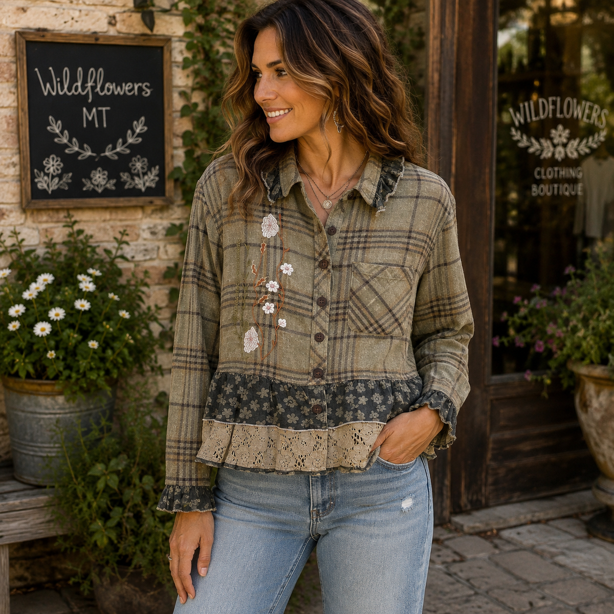 A woman wearing a long-sleeve plaid button-up shirt with floral embroidery and lace trim, paired with light-wash blue jeans, standing in front of a rustic storefront with a chalkboard sign reading Wildflowers MT.