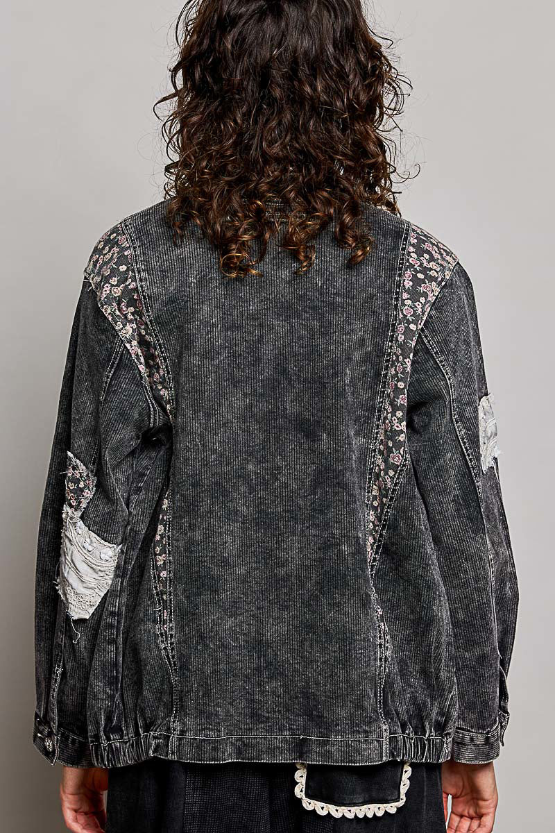 A person shown from the back wearing a washed black denim jacket with floral lace applique along the shoulders and sleeves, paired with dark pants against a plain light gray studio background.