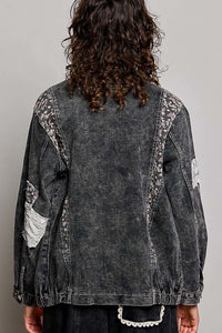 A person shown from the back wearing a washed black denim jacket with floral lace applique along the shoulders and sleeves, paired with dark pants against a plain light gray studio background.