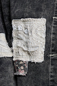 Close-up of a dark charcoal ribbed fabric garment featuring a distressed patch pocket with layered off-white lace applique and a small floral fabric tab.