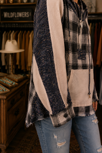 A person wearing a patchwork waffle-knit hoodie with navy, black, and beige panels, paired with distressed blue jeans in a rustic clothing store.