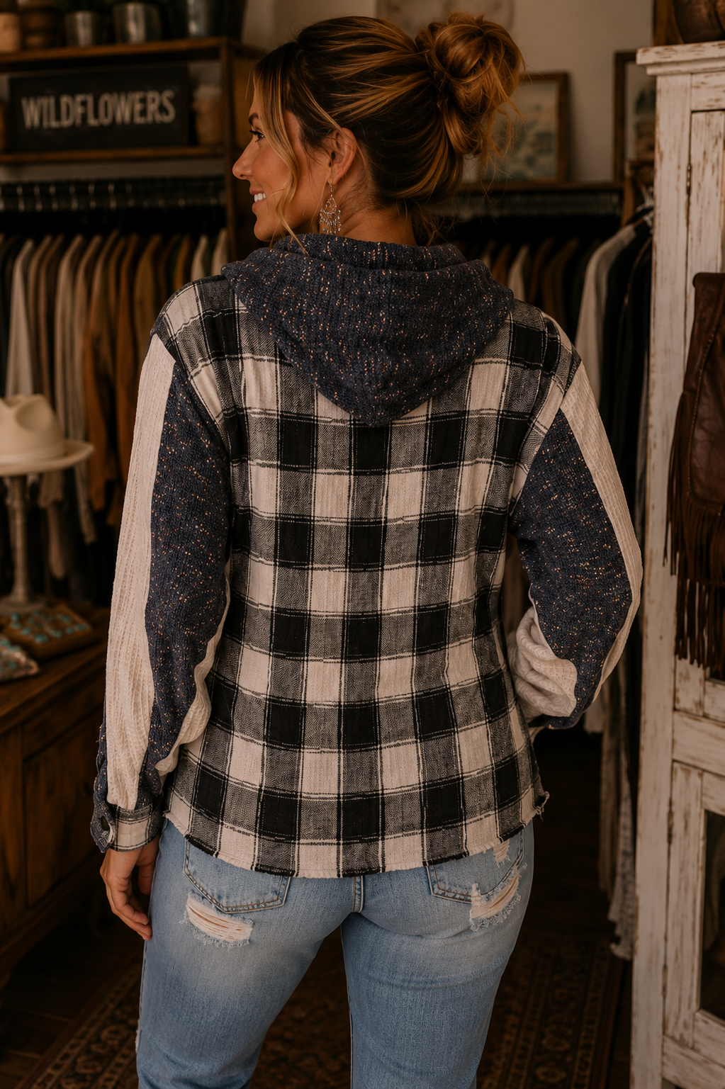A woman shown from the back wearing a hooded plaid jacket with a black and white buffalo check pattern and textured navy sleeves, standing inside a rustic clothing store.
