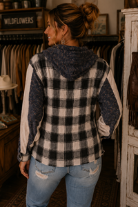 A woman shown from the back wearing a hooded plaid jacket with a black and white buffalo check pattern and textured navy sleeves, standing inside a rustic clothing store.