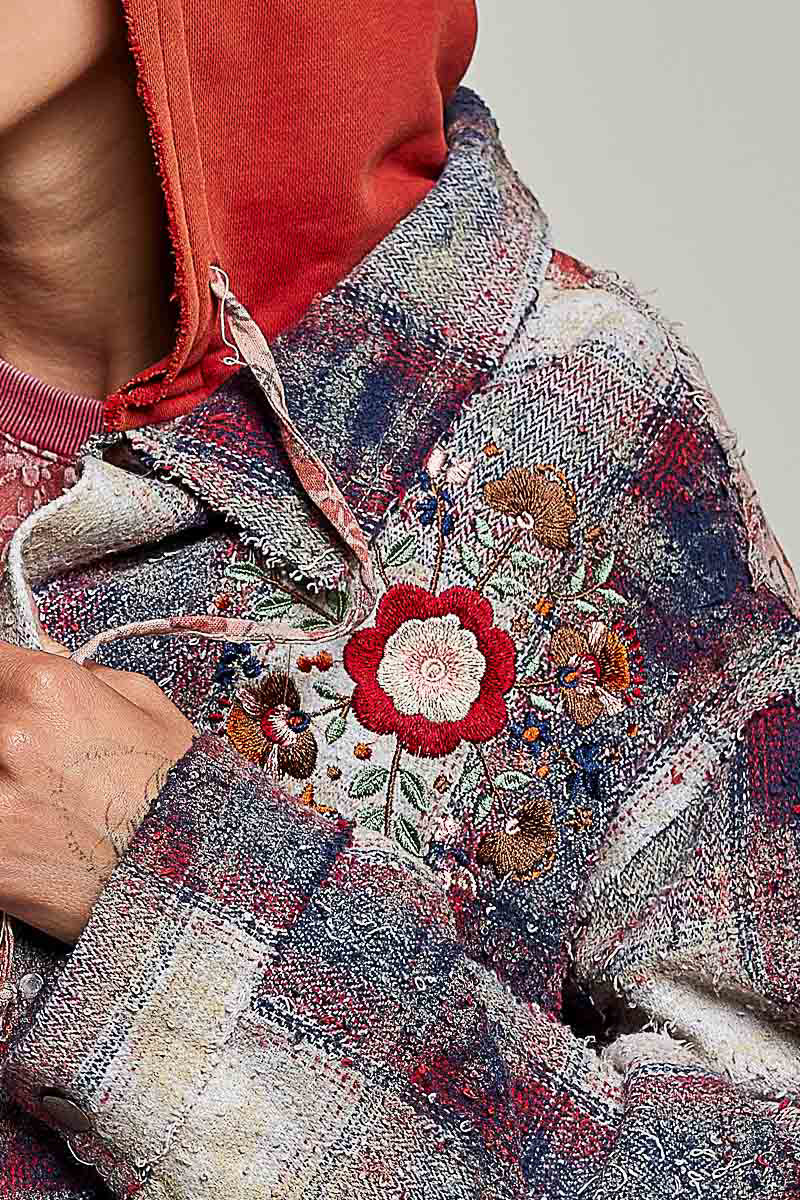 Close-up of a person wearing a heavily textured, frayed patchwork jacket featuring a red rose embroidery on the chest, layered over a red hooded sweatshirt.