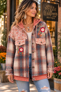 A woman wearing a hooded plaid jacket with floral patches and a pink hood, standing outdoors in front of a boutique storefront.