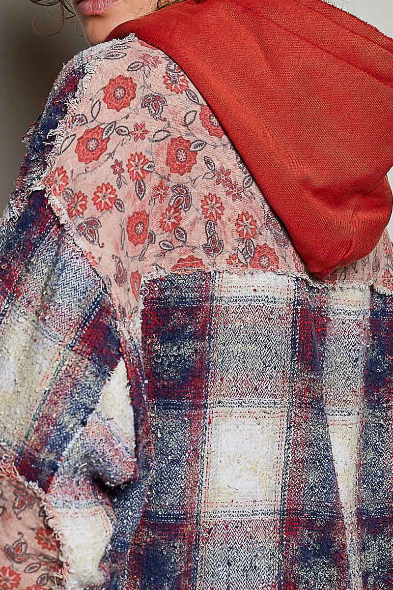 Close-up of a person wearing a patchwork hoodie with a red hood, a beige floral panel, and a distressed plaid patch in blue, red, and cream.