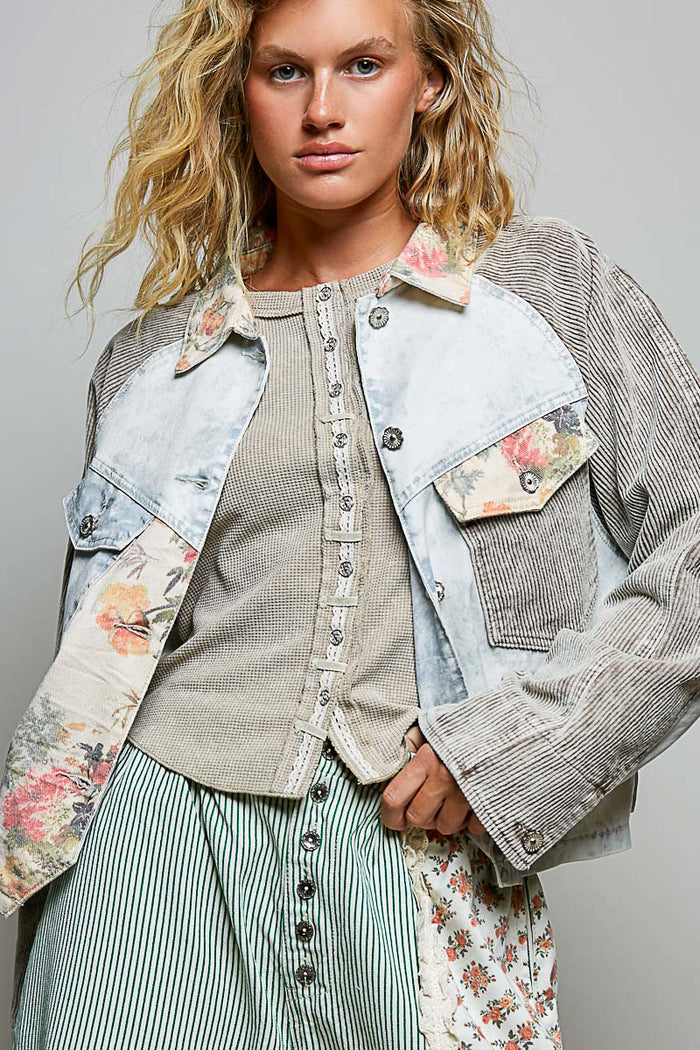 A woman wearing a layered fashion outfit featuring a light-wash denim jacket with floral printed panels, a ribbed beige top, and a green-and-white striped skirt, photographed against a neutral gray studio background.