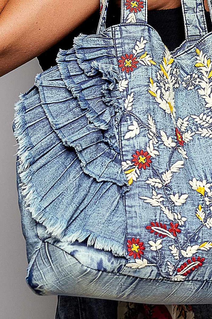 A close-up of a denim tote bag being carried on a person's shoulder. The bag has layered ruffle details, frayed edges, and colorful floral embroidery in red, yellow, and white on a light blue denim background.