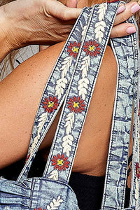 Close-up of a person holding a distressed denim bag with wide embroidered shoulder straps featuring red flowers, yellow centers, and white leaf patterns.
