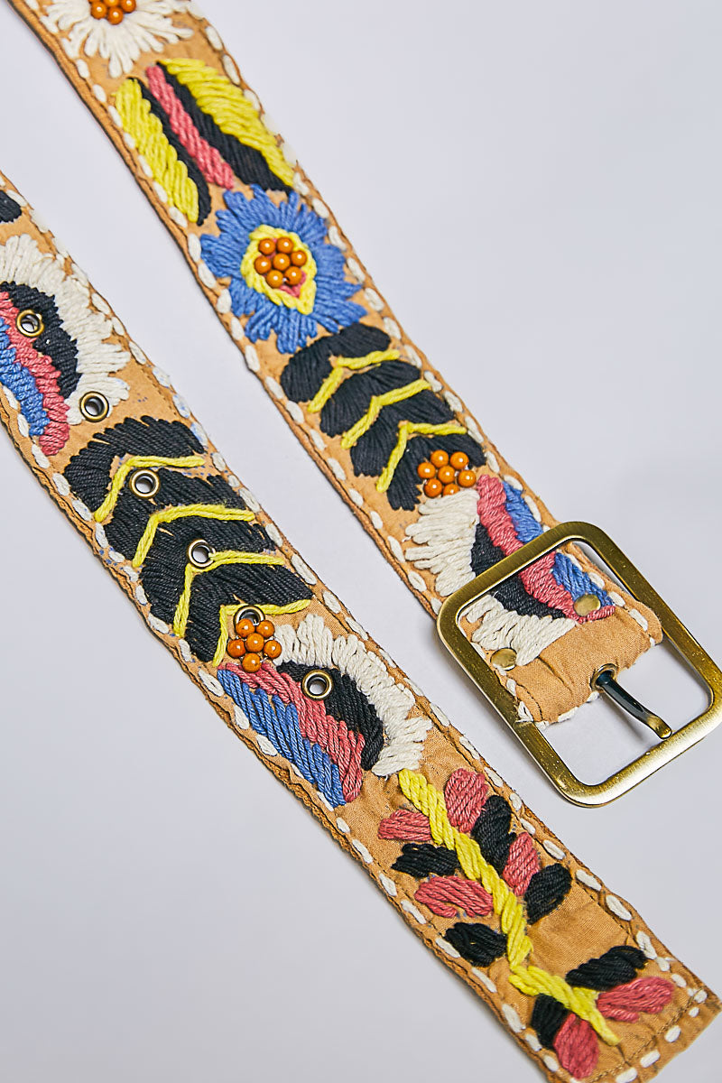 A close-up of a tan leather belt with colorful embroidered floral and geometric patterns, featuring a gold-tone metal buckle, set against a light gray background.