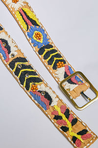 A close-up of a tan leather belt with colorful embroidered floral and geometric patterns, featuring a gold-tone metal buckle, set against a light gray background.