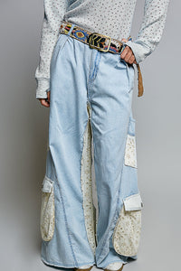 A person wearing light blue wide-leg denim cargo pants with cream patch pockets and a colorful patterned belt, paired with a white patterned long-sleeve top against a gray studio background.