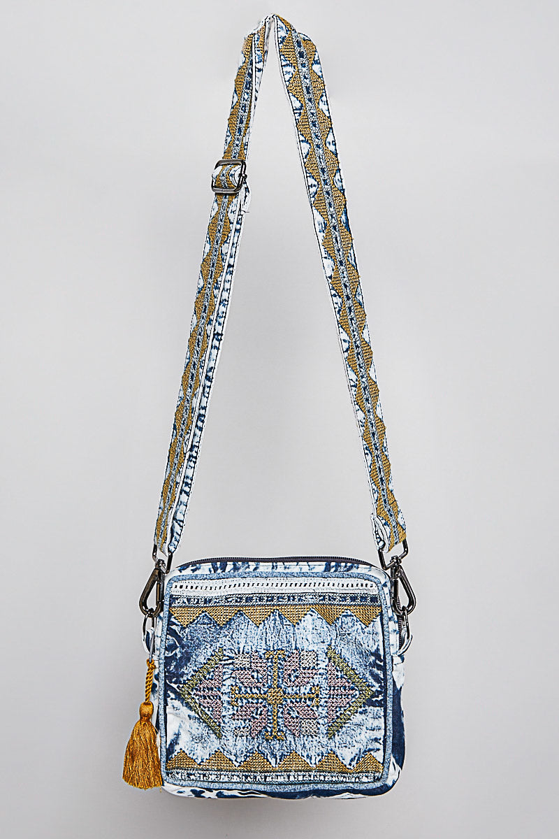 A small crossbody bag with a blue, white, and gold geometric pattern, featuring a matching patterned shoulder strap and a gold tassel, shown against a light gray background.