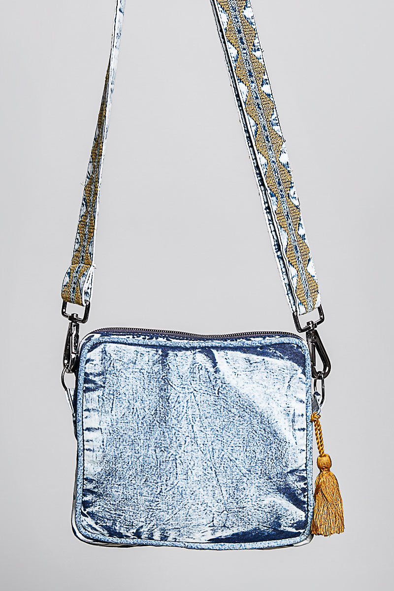 A washed crossbody bag, featuring a patterned strap with gold and silver details and a gold tassel charm, shown against a plain light gray background.