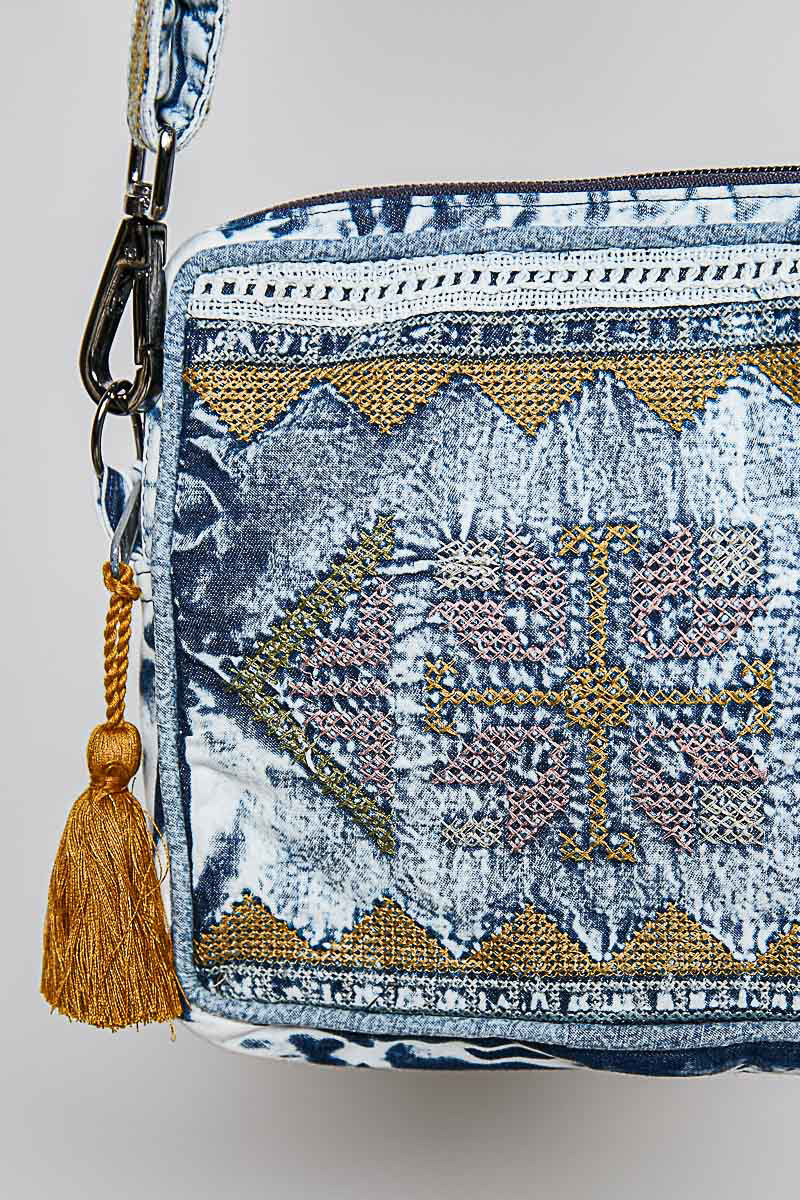 A close-up of a patterned crossbody bag featuring a blue and white base with gold, pink, and blue geometric embroidery and a gold tassel charm.