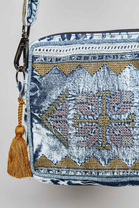 A close-up of a patterned crossbody bag featuring a blue and white base with gold, pink, and blue geometric embroidery and a gold tassel charm.