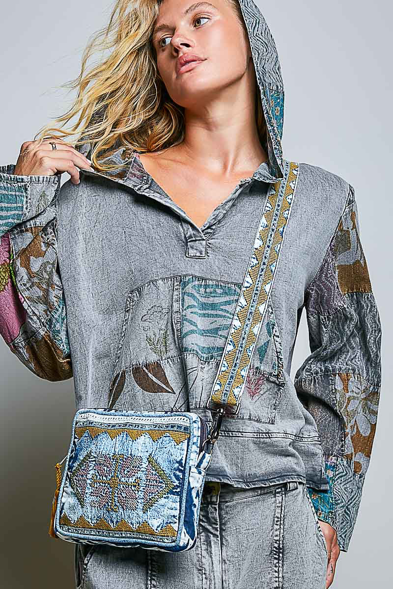 A woman wearing a gray hooded patchwork top with colorful embroidered details and a matching denim embroidered crossbody bag, shown against a light gray studio background.