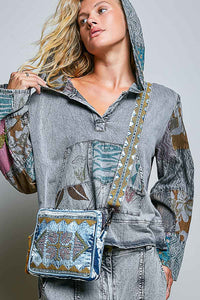 A woman wearing a gray hooded patchwork top with colorful embroidered details and a matching denim embroidered crossbody bag, shown against a light gray studio background.