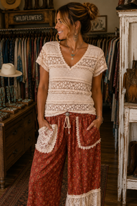 A woman modeling a bohemian-style two-piece outfit in a rustic clothing boutique. The set features a cream lace short-sleeve top and rust-red patterned wide-leg pants with lace trim.