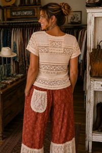 A woman stands in a rustic clothing boutique wearing a cream lace-trimmed short-sleeve top and rust-red patterned wide-leg pants with lace hem and pocket details.