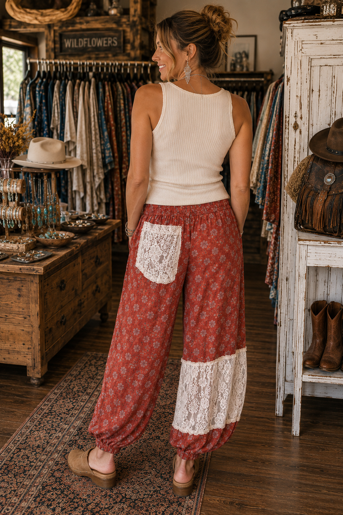 A woman stands in a rustic clothing boutique wearing loose, patterned rust-red harem-style pants with white lace pocket and lower-leg panels, paired with a cream ribbed tank top.