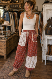 A woman stands in a rustic clothing boutique wearing loose, flowy patterned pants with white lace patch pockets and a white ribbed tank top. The pants are rust red with a small floral print and white lace sections near the hems.