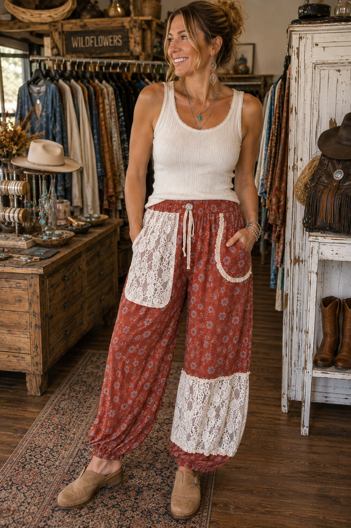 A woman stands in a rustic clothing boutique wearing loose, flowy patterned pants with white lace patch pockets and a white ribbed tank top. The pants are rust red with a small floral print and white lace sections near the hems.