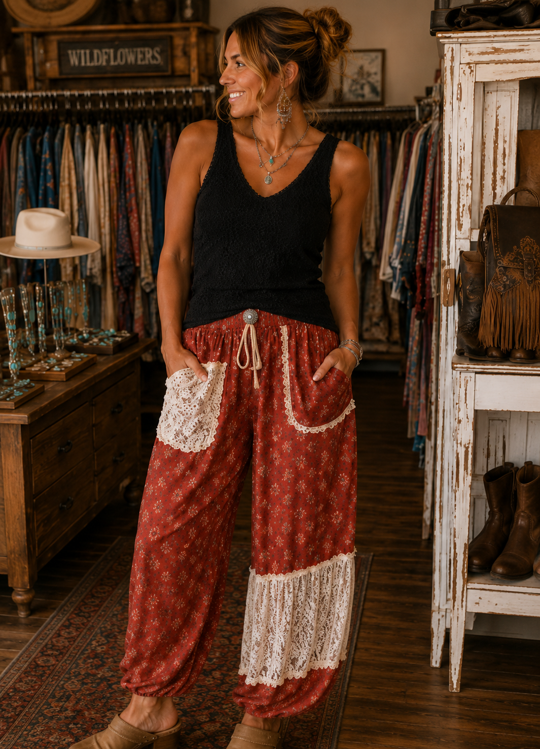 A woman stands in a rustic clothing boutique wearing loose red patterned pants with white lace trim and a black sleeveless top. The boutique has wooden racks of colorful garments, a white cabinet, and brown boots on display.