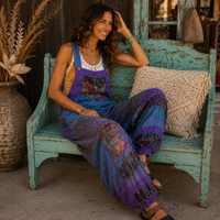 A woman sits on a distressed turquoise wooden bench wearing a colorful patchwork jumpsuit with purple, blue, and orange tones. The setting is rustic with a large ceramic vase of dried grass, a crocheted pillow, and a dark wooden wall in the background.