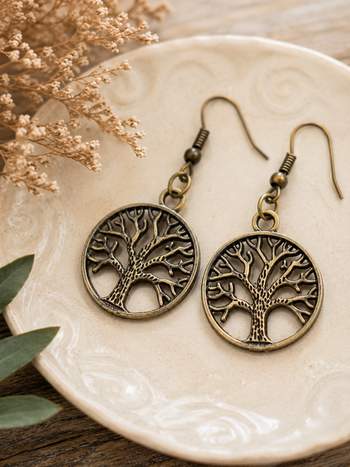 A pair of antique bronze tree-of-life drop earrings displayed on a beige ceramic plate with dried flowers and green leaves.