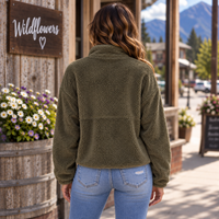 A woman viewed from behind wearing an olive green sherpa fleece jacket and light blue jeans, standing on a street in front of a wooden building with a sign that reads 'Wildflowers'.
