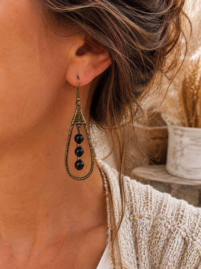 A close-up of a woman wearing a single dangling earring. The earring has an antique bronze-toned metal frame with three dark, nearly black round beads hanging in the center.