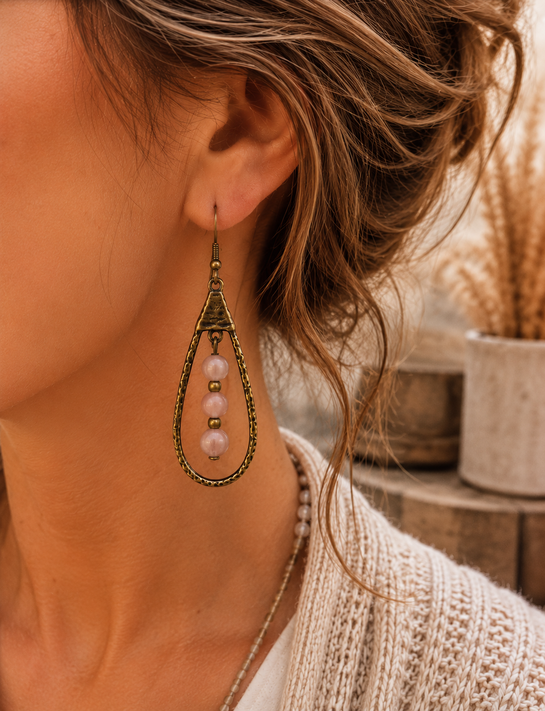 A close-up lifestyle shot of a woman wearing a single teardrop-shaped dangling earring. The earring features an antique bronze textured frame with three light pink pearl-like beads hanging in the center.