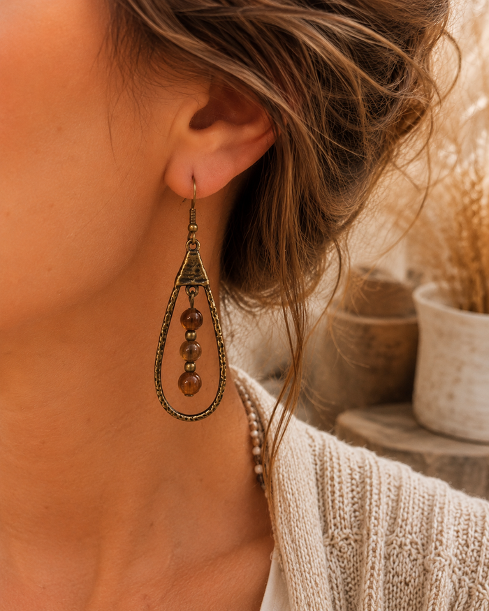 A close-up of a woman wearing a long, ornate teardrop dangle earring with bronze-toned metalwork and amber-brown beads, shown against a softly blurred boho-style background.