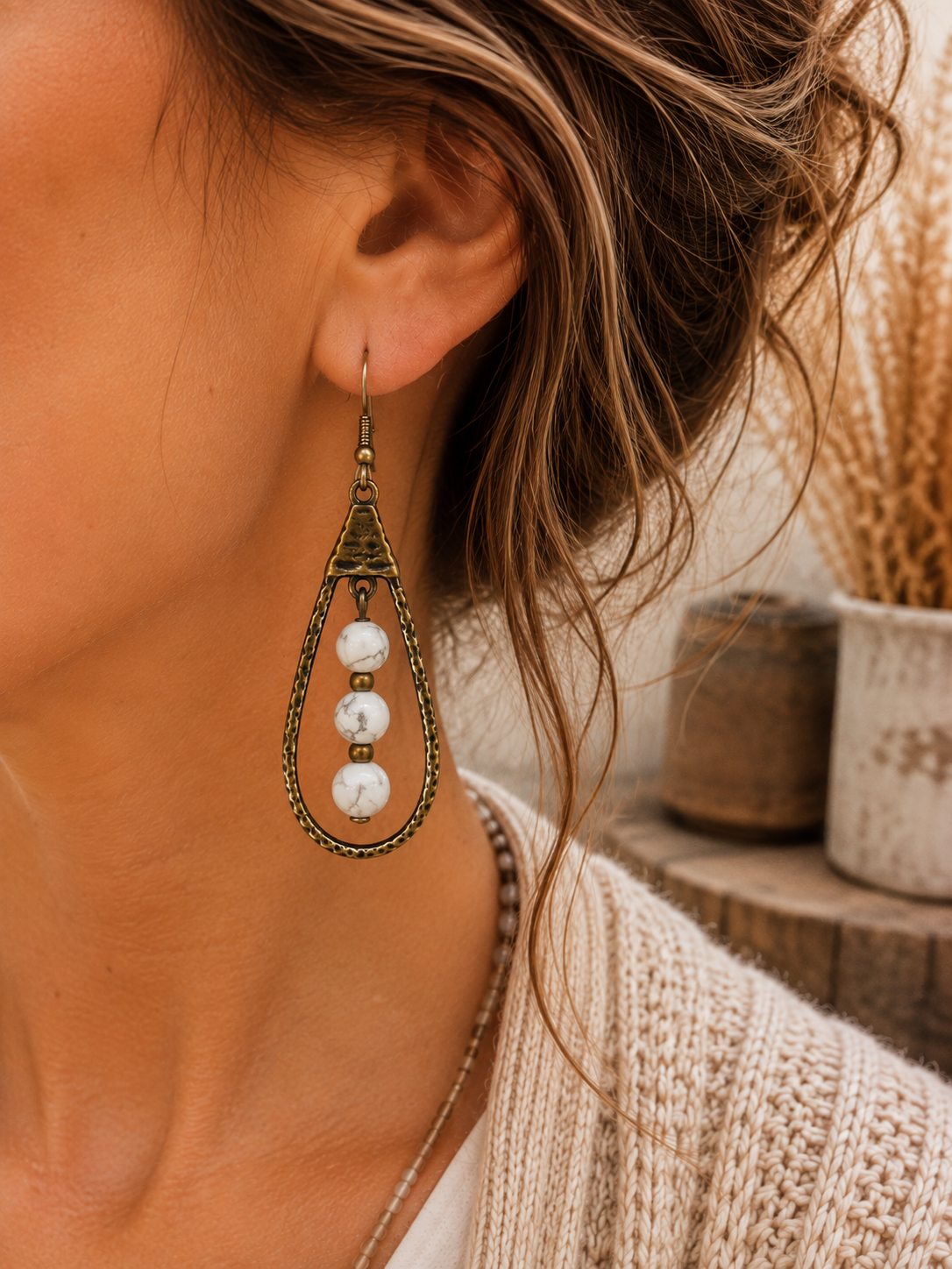 A close-up lifestyle shot of a woman wearing a dangling teardrop earring. The earring features an antique bronze metal frame with three white marbled beads.