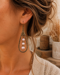 A close-up of a woman wearing a long teardrop-shaped dangling earring with an antique bronze metal frame and three pink pearl beads hanging down.